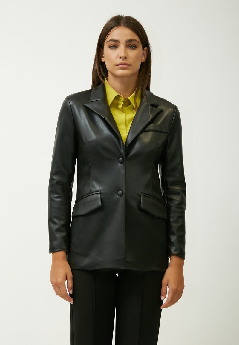 Blazer in pelle nera con design aderente, chiusura a bottone singolo, due tasche frontali e una texture liscia. Indossato sopra una camicia gialla brillante.