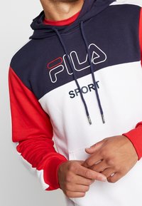 Sudadera con capucha en azul marino, rojo y blanco. Presenta un logo en el pecho, capucha con cordones y puños acanalados. Confeccionada en una tela suave con un diseño deportivo.