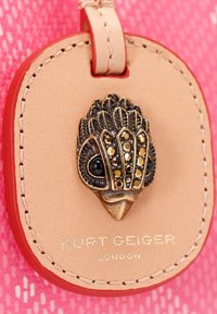 Etichetta in pelle con un elegante accento a forma di uccello color oro, impreziosita da gemme nere. La scritta riporta "Kurt Geiger London." L'etichetta presenta uno sfondo rosa e rosso.