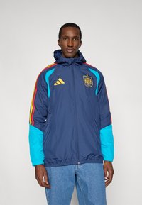 Marineblauwe windbreaker met capuchon; heeft turquoise en rode accenten, het Adidas-logo en het embleem van het Spaanse nationale team op de borst. Lichtgewicht stof.