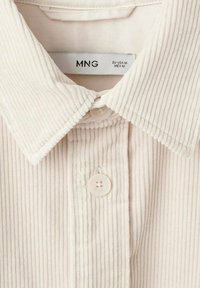 Camisa de pana crema con cuello de solapa, dos botones y textura de ribetes verticales. La etiqueta muestra la marca y detalles de tamaño.