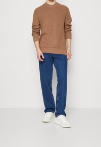 Brązowy sweter z prążkowanego materiału z okrągłym dekoltem, w zestawie z niebieskimi prostymi dżinsami i białymi trampkami. Prosty design, swobodny krój.