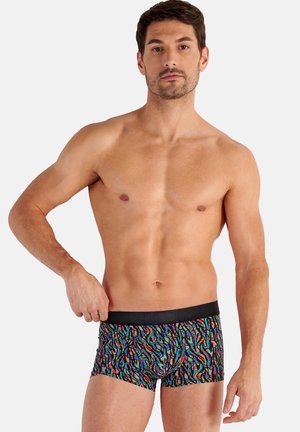 Mann mit kurzen dunklen Haaren steht oben ohne und trägt bunte gemusterte Boxershorts mit schwarzem Bund vor weißem Hintergrund.