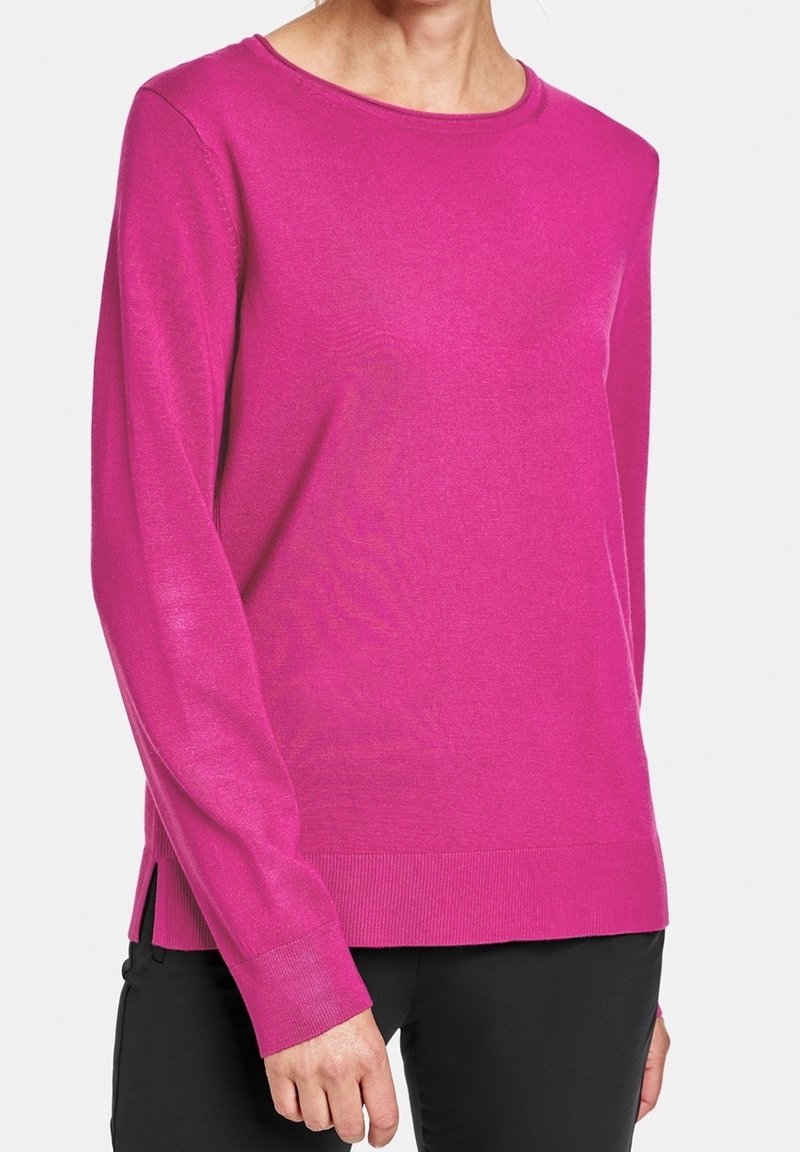 Kobieta ubrana w jasnoróżowy sweter z długim rękawem i bocznymi rozcięciami, w połączeniu z czarnymi spodniami, stojąca na tle białym.