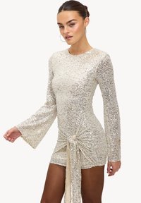 Robe en sequins argentés avec de longues manches évasées, un col rond et un détail noué à l'avant. Texture lisse et silhouette ajustée.