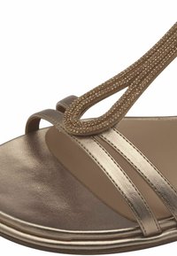 Sandale dorée métallique avec deux brides étroites et un détail de T-bride décoré de bijoux. Texture en cuir lisse et semelle intérieure rembourrée.