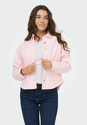 Femme portant une veste rose clair boutonnée sur un haut blanc et un jean bleu foncé, ajustant la veste avec les deux mains.