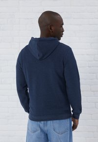 Marineblauwe hoodie met lange mouwen, textuurstof en een kangoeroezak, gedragen met lichtblauwe jeans tegen een witte bakstenen muur.
