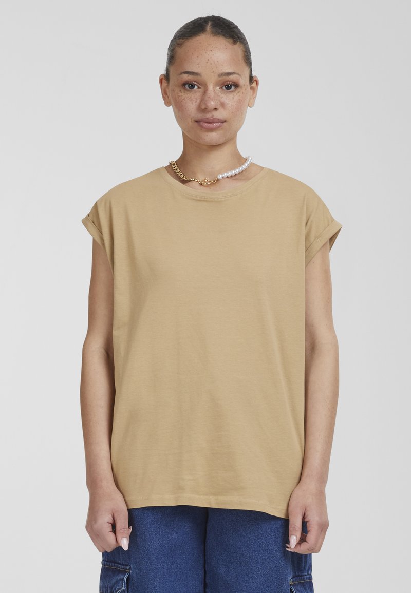 Beige, oversize tričko s krátkými rukávy na ohrnutí. Jemná bavlněná tkanina, kulatý výstřih. Nosené s denimovými spodky a vícero vrstvenými náhrdelníky.