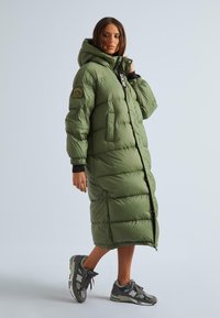 Olive zaļš pūkains mētelis ar garu siluetu, kapuci un spiedpoga aizdari. Iezīmēts ar izšūtu šuvju rakstu un logo uz piedurknes.