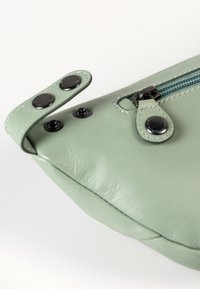 Groene leren tas met een gladde textuur, uitgerust met zwarte drukknoopjes en een contrasterende blauwe rits, die de vorm en functionaliteit accentueert.