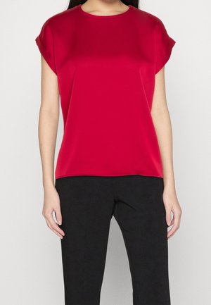 Femme portant un chemisier rouge vif à manches courtes avec des manches retroussées et un pantalon noir sur un fond uni.