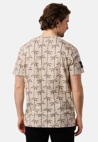 Beige cotton t-shirt featuring a repeating palm tree pattern in dark brown. Classic fit with a crew neck and short sleeves.