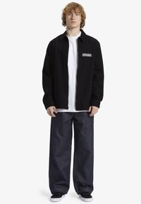 Chaqueta negra con cremallera sobre una camiseta blanca, combinada con unos jeans de mezclilla oscuros de pierna ancha y zapatillas negras, que presentan un detalle de logo en la chaqueta.