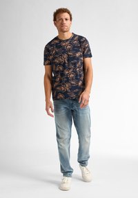 Camiseta floral azul marino con patrones beige, combinada con jeans de un azul claro y zapatillas blancas. La camiseta tiene un cuello redondo y mangas cortas.