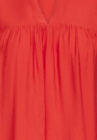 Abito rosso con scollatura a V, caratterizzato da pieghe arricciate sul seno. Il tessuto ha una finitura morbida e testurizzata, con una silhouette fluida.