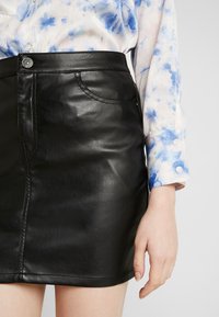 Černá kožená mini sukně s hladkým povrchem, s přední kapsou a kovovým zapínáním. Kombinováno s košilí s modrým tie-dye vzorem.