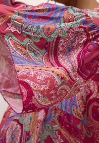 Kolorowa tkanina w wzór paisley w odcieniach różu, zieleni i niebieskiego. Miękka w dotyku, z falbaniastymi brzegami i skomplikowanymi detalami na całej powierzchni.