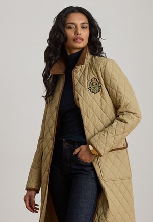 Donna con un lungo cappotto trapuntato beige con stemma ricamato, dolcevita scuro e jeans scuri, in piedi con una mano in tasca.