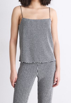 Kobieta nosząca dopasowany czarno-biały, pionowo prążkowany, bezrękawnik z cienkimi ramiączkami i spodnie na jednolitym tle.