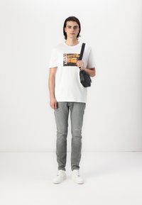 T-shirt blanc avec imprimé graphique, jeans gris déchirés et sac bandoulière noir. Baskets blanches avec semelles texturées.