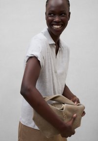 Personne souriante portant une chemise blanche à manches courtes et tenant un sac en tissu beige plié sur un fond clair uni.