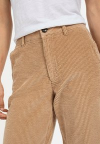 Brune fløjlshorts med lodrette ribber, med standard knaplukning og bæltesløjfer. Nærbillede af tekstureret stof.