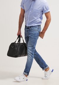 Musta nahkainen duffel-laukku, jossa on kahdet kahvat, kannettuna henkilö toimissa vaaleansininen ruutupaita, siniset farkut ja valkoiset tennarit sinisin yksityiskohdin.