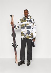 Hav snowboardos, amely álcázó dzsekit visel fehér, szürke és zöld mintázattal. Fekete nadrág, sícipők, és egyik kezében síléceket tart.