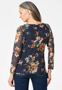 Blumiges Langarmoberteil aus transparentem schwarzem Stoff mit lebhaften Blumenmustern in Orange, Blau und Pink; runder Halsauschnitt, eng anliegende Ärmel, hüftlang.