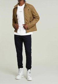 Beige zipjacka med hög krage, vit t-shirt med rund hals under, mörka byxor och vita snörstövlar, med en logotypdetalj.