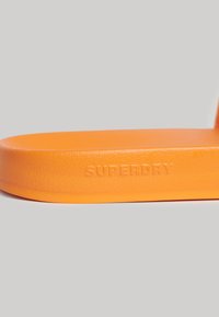 Oranje rubberen sandal met een gestructureerde oppervlakte en een verhoogd "SUPERDRY" logo. De zool heeft een gladde afwerking, die de levendige kleur benadrukt.