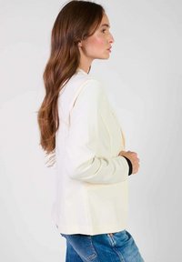 Blazer couleur crème avec une coupe ajustée, dotée d'un col cranté, de poches latérales et d'accents noirs contrastants au niveau des poignets.