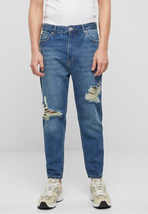 Jean en denim bleu avec une coupe décontractée, présentant des déchirures usées au niveau des genoux et un léger dégradé. Le tissu semble doux avec une texture lisse.
