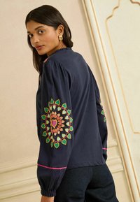 Marineblauwe geborduurde blouse met opgeblazen mouwen, voorzien van veelkleurige hartpatronen op de armen en roze bies bij de manchetten.