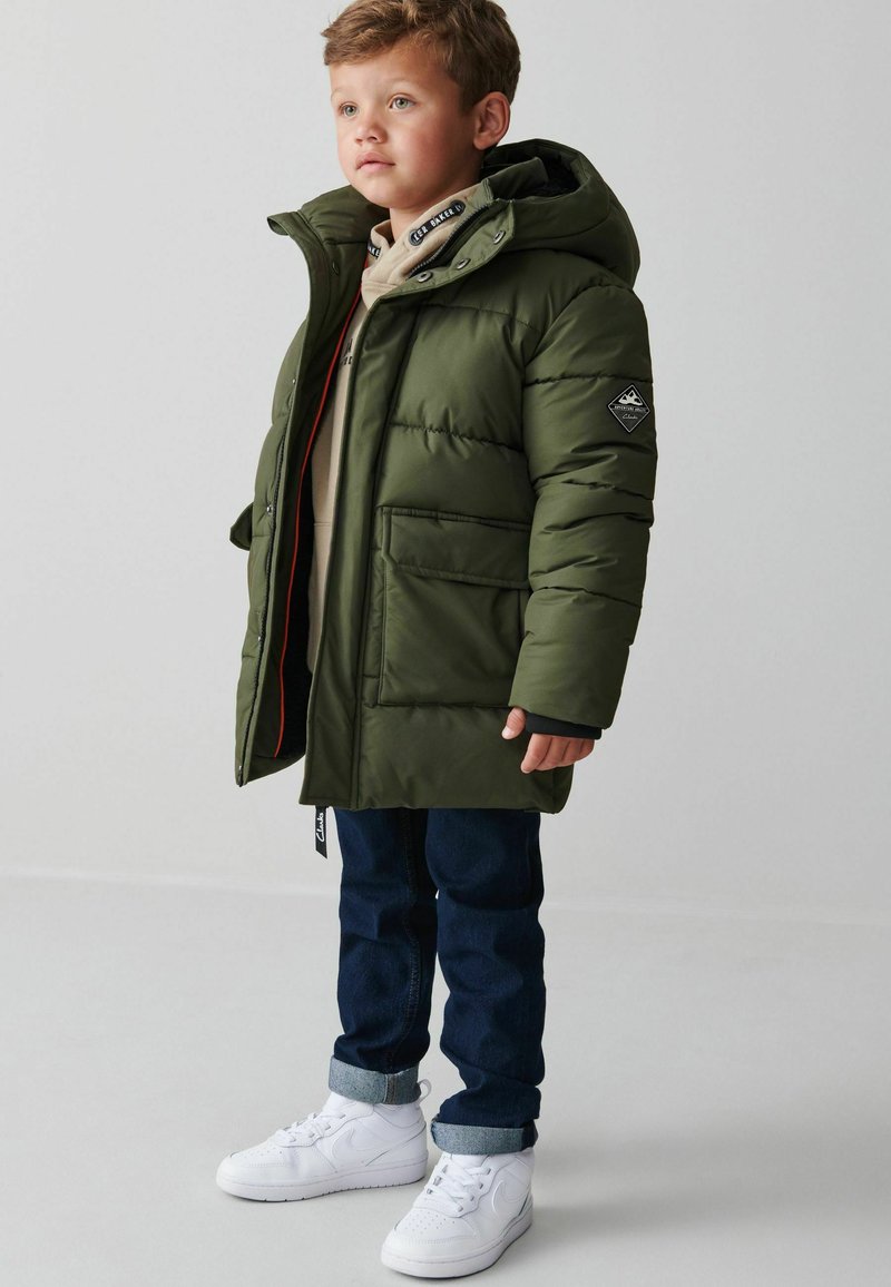 Giacca imbottita verde con cappuccio, tasche frontali e un patch con logo sulla manica. Indossata sopra abiti beige e abbinata a jeans scuri e sneakers bianche.