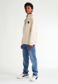 Felpa beige con zip al collo e toppa con logo nero, abbinata a jeans di denim blu e sneakers bianche con dettagli neri. Tessuto dalla finitura liscia.