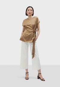 Top in satin marrone con vita annodata, abbinato a pantaloni bianchi a gamba larga e sandali neri con tacco blocco e un accento decorativo.
