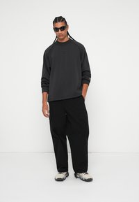 Melna pārāk liela izmēra sporta krekls ar apaļu kakla izgriezumu, savienots ar melniem biksēm ar kabatām. Modelis valkā tumšas saulesbrilles un bēšas sporta apavus.