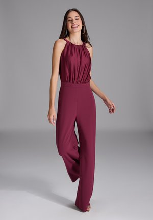 Femme souriante portant une combinaison bordeaux sans manches avec un col montant froncé et un pantalon à jambes larges, debout sur un fond gris.