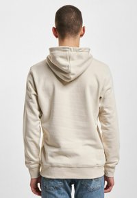 Beige Kapuzenpullover aus einem weichen, strukturierten Stoff. Elastische Bündchen und Saum, minimalistisches Design und eine Kängurutasche vorne.