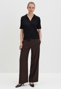 Zwarte ribbelige top met korte mouwen en V-hals, gecombineerd met lange bruine broeken en zwarte puntige schoenen. Eenvoudig, gestroomlijnd ontwerp.