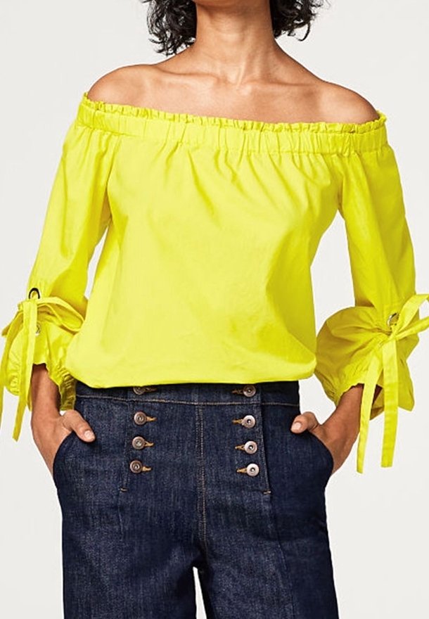 Femme portant un chemisier jaune vif à épaules dénudées avec des manches 3/4 nouées et un pantalon bleu foncé taille haute avec deux rangées de boutons.