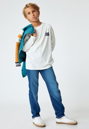 Wit longsleeve shirt met een logo op de borst, blauwe denim jeans, een teal en oranje jas, en witte sneakers met beige accenten.