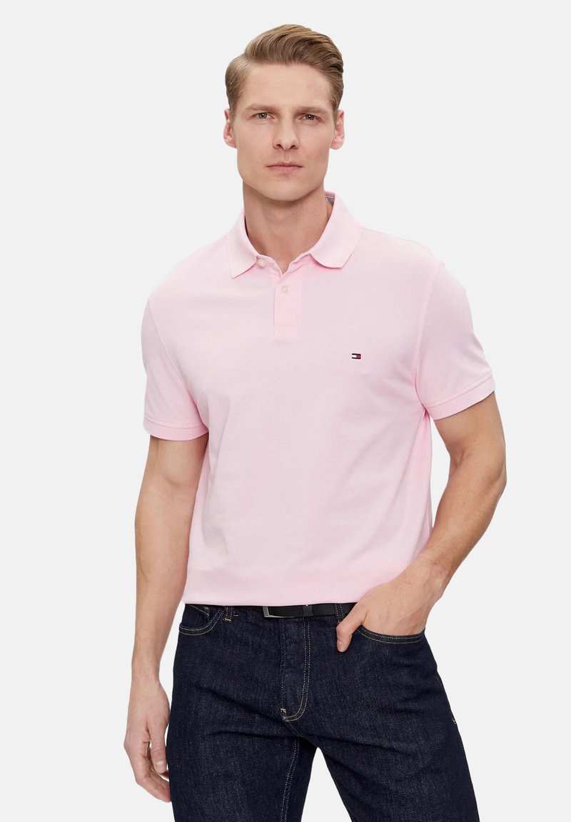 Polo de color rosa claro hecho de algodón, con una tapeta de dos botones, mangas cortas y un pequeño logo en el pecho. Combinado con jeans oscuros.