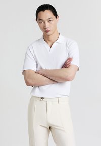 Polo bianco a righe con maniche corte, caratterizzato da un colletto a V. Abbinato a pantaloni beige chiaro, il modello sta in piedi con le braccia incrociate.