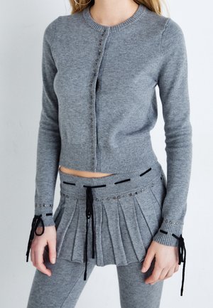 Vrouw draagt een grijs gebreid vest met lange mouwen en bijpassende geplooide rok met zwarte kanten details en leggings tegen een effen achtergrond.