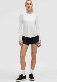 Bijela majica dugih rukava za sport s prozirnim ramenima, uparena s crnim šortsevima i crnim tenisicama. Jednostavan, funkcionalan dizajn.