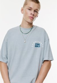 Ung mand med kort blond hår iført en lysblå "STWD GOALS" t-shirt og to halskæder, poserer mod en ensartet hvid baggrund.