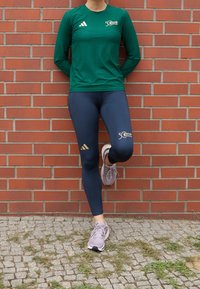 Långärmad grön sporttröja med en logotyp, kombinerad med marinblå leggings med guld accents och en logotyp. Bärs med ljuslila sneakers.